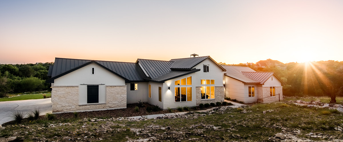 3911 Exterior view of modern farmhouse at sunset with large windows, stone veneer, and metal roof.