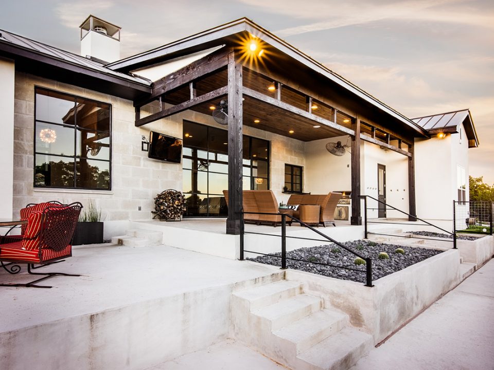 Modern farmhouse back patio with wood beam pergola, stone exterior, and outdoor seating area at sunset.
