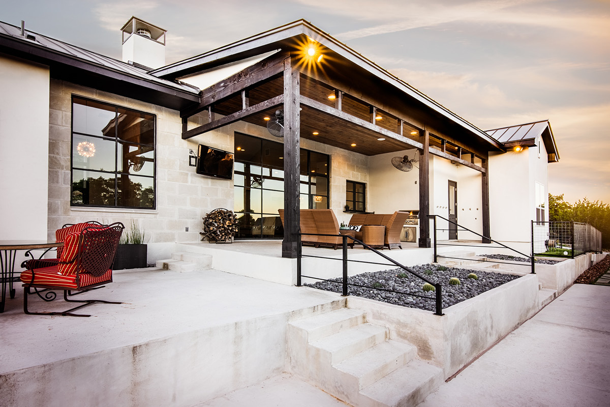 3911-8 Modern farmhouse back patio with wood beam pergola, stone exterior, and outdoor seating area at sunset.