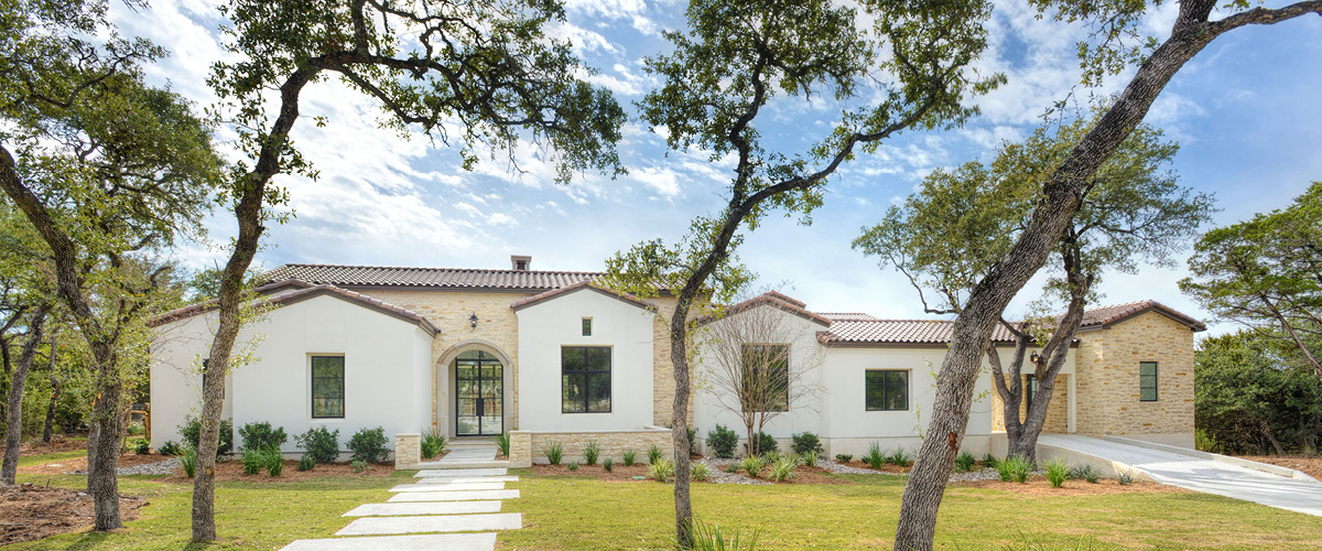Spanish Revival home exterior with clay tile roof, white stucco walls, arched front entry, and landscaped front yard with native trees.