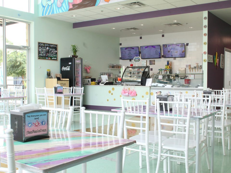 Whimsical ice cream shop interior at Bee’s Neez Cones & Coffee in San Antonio featuring pastel colors, white chairs, glitter floor, and ice cream counter with menu screens.