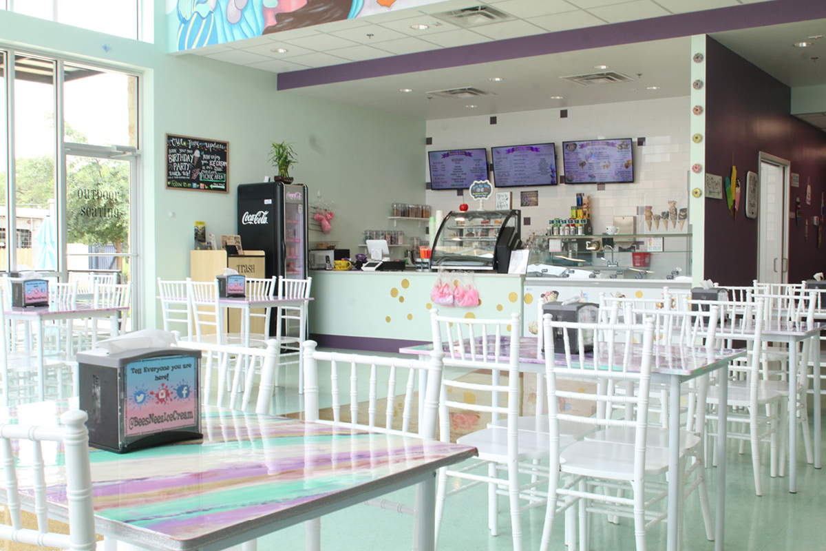 Whimsical ice cream shop interior at Bee’s Neez Cones & Coffee in San Antonio featuring pastel colors, white chairs, glitter floor, and ice cream counter with menu screens.