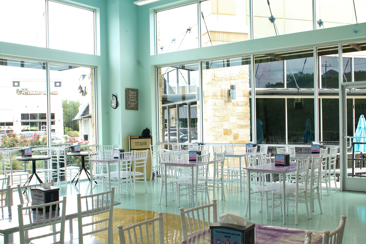 Bright dining area at Bee’s Neez Cones & Coffee in San Antonio with floor-to-ceiling windows, white tables and chairs, and cheerful pastel walls.