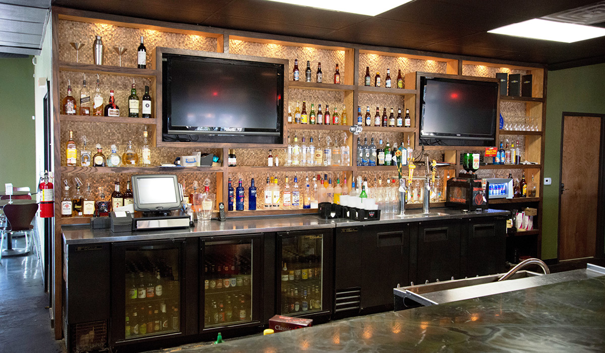 evil olive -2 Close-up view of Evil Olive’s bar with liquor shelves, mounted TVs, and illuminated shelving.