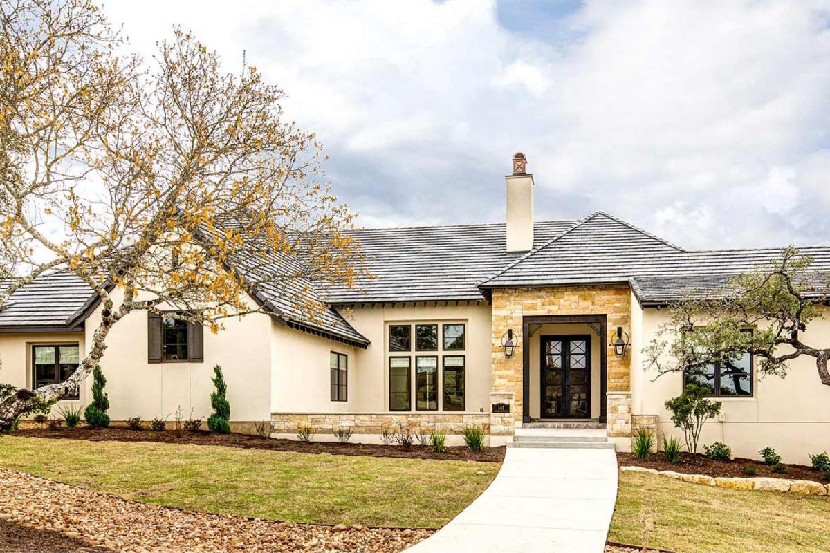 4880- French country home exterior with stone entryway, tall windows, and a curved walkway framed by oak trees.