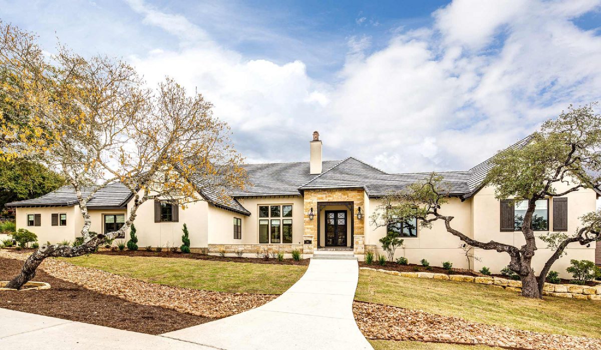 4880-2 French country style home exterior with light stone accents, arched entry, and mature oak trees framing the front walkway.