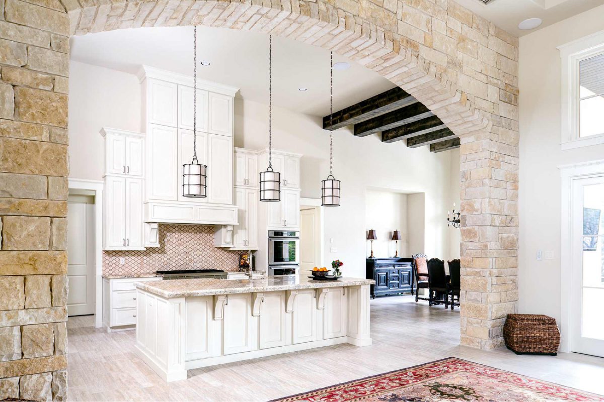 4880-4 French country kitchen with stone archway, white cabinetry, large island, and pendant lights opening into dining area with exposed ceiling beams