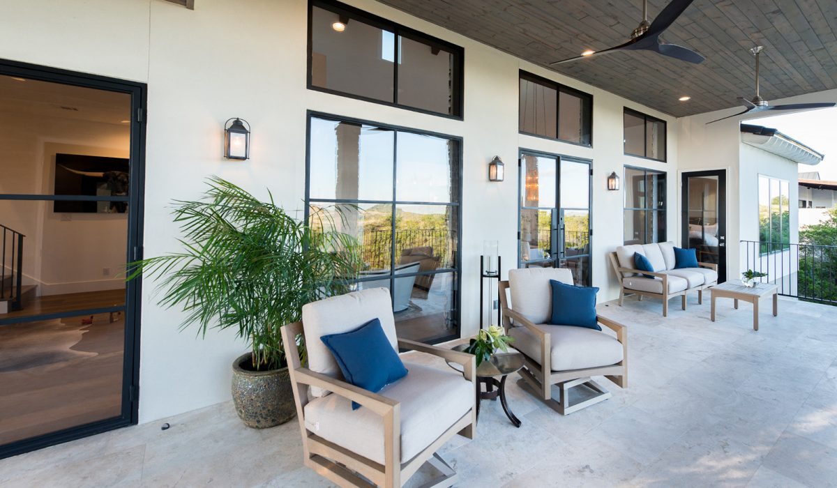 3674-16 Elegant stone-tiled patio featuring cushioned lounge chairs, blue accent pillows, potted greenery, and black-framed windows overlooking scenic hill country.