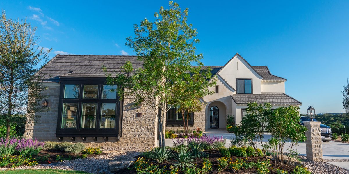 Elegant stone and stucco home with arched entryway, tall windows, and landscaped front yard under a clear blue sky