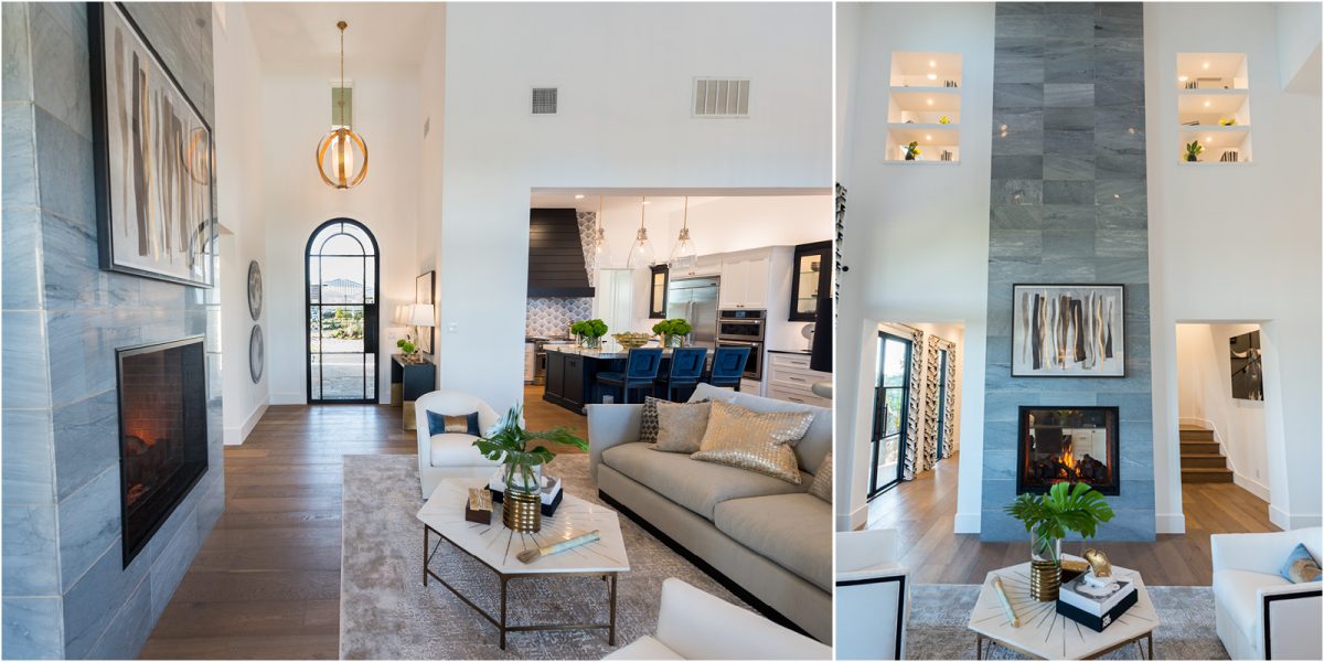 Bright living room with a tall stone fireplace, modern chandelier, and open layout connecting to the kitchen with blue barstools.